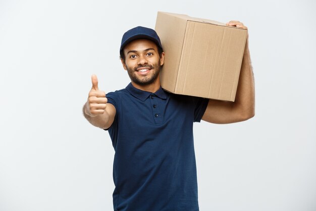 Conceito de entrega - Retrato de um homem de entrega feliz americano africano apontando mão para apresentar um pacote de caixa. Isolado no fundo do estúdio cinzento. Espaço de cópia.
