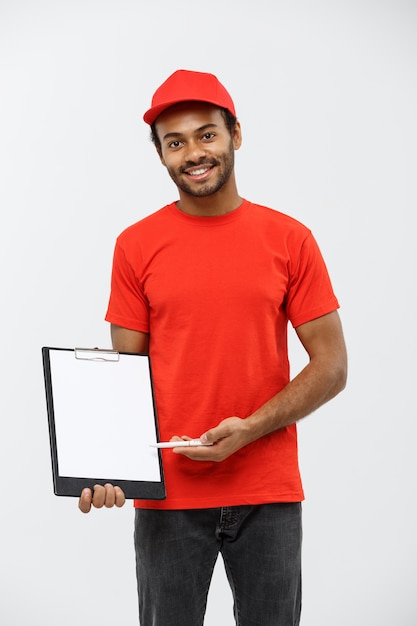 Conceito de entrega - Retrato do homem de entrega afro-americano bonito ou de um mensageiro que mostra um formulário de documento de confirmação para assinar. Isolado no fundo do estúdio cinzento. Espaço de cópia.