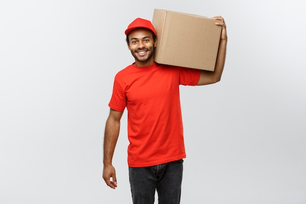 Conceito de entrega - Retrato do homem de entrega feliz Africano-americano em pano vermelho, segurando um pacote de caixa. Isolado no fundo do estúdio cinzento. Espaço de cópia.
