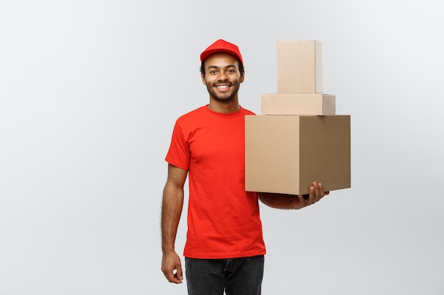 Conceito de entrega - Retrato do homem de entrega feliz Africano-americano em pano vermelho, segurando um pacote de caixa. Isolado no fundo do estúdio cinzento. Espaço de cópia.