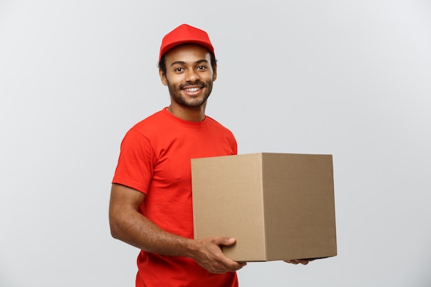 Conceito de entrega - Retrato do homem de entrega feliz Africano-americano em pano vermelho, segurando um pacote de caixa. Isolado no fundo do estúdio cinzento. Espaço de cópia.