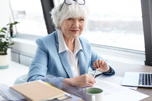 Conceito de envelhecimento, aposentadoria, carreira e emprego. Retrato de uma atraente CEO feminina, branca, na casa dos sessenta anos, trabalhando na mesa em frente a um computador aberto, sentada perto da janela, desfrutando de sua ocupação