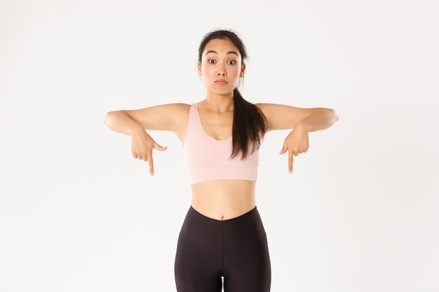 Conceito de esporte, bem-estar e estilo de vida ativo. Menina asiática de fitness assustada e impressionada, membro da academia ou desportista em roupa ativa, apontando os dedos para baixo, olhando sem palavras, fundo branco.