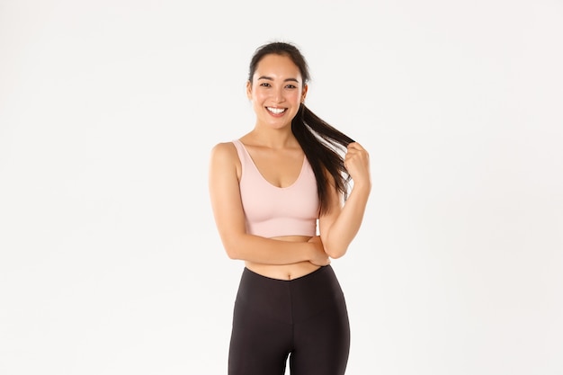 Conceito de esporte, bem-estar e estilo de vida ativo. Menina muito asiática no sportswear, brincando com um rabo de cavalo e olhando feliz para a câmera após um bom treino de ginástica, fundo branco de pé a sorrir.