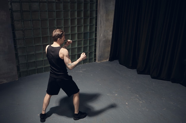 Conceito de esportes, fitness e determinação. vista traseira de um jovem e musculoso kickboxer masculino de tênis preto, shorts e camiseta regata trabalhando em socos em uma sala vazia, segurando os punhos na frente dele