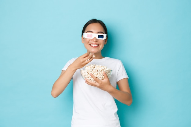Conceito de estilo de vida, lazer e emoções. Sorrindo satisfeita menina asiática parecendo satisfeita enquanto come pipoca da tigela, assistindo filme na tela de TV com óculos 3d, desfrutando de uma série incrível.