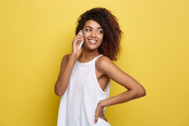 Conceito de estilo de vida - Retrato da bela mulher afro-americana alegre conversando no celular com amigo. Fundo de estúdio amarelo pastel. Espaço de cópia.