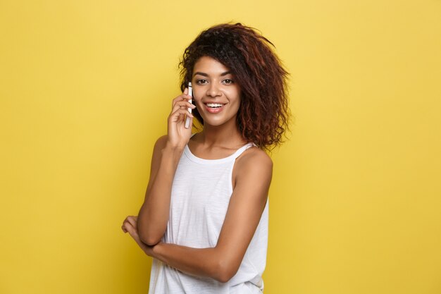 Conceito de estilo de vida - Retrato da bela mulher afro-americana alegre conversando no celular com amigo. Fundo de estúdio amarelo pastel. Espaço de cópia.