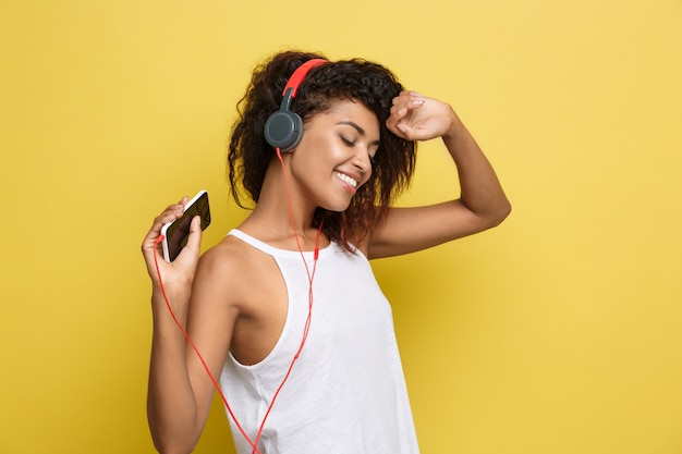 Foto grátis conceito de estilo de vida - retrato da bela mulher afro-americana alegre, ouvindo música no celular. fundo de estúdio amarelo pastel. espaço de cópia.