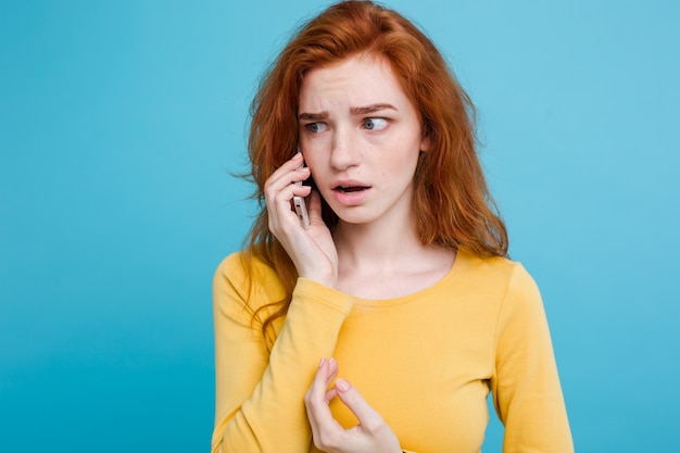 Conceito de estilo de vida - retrato de menina de cabelo vermelho gengibre com expressão chocante e estressante enquanto conversa com um amigo por telefone celular. isolado no fundo do pastel azul. copie o espaço.