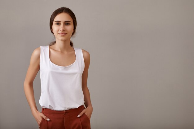 Conceito de estilo, moda, elegância e confiança. Jovem europeia elegante com ombros nus e bronzeados posando isolada contra a parede vazia do copyspace, mantendo as mãos nos bolsos