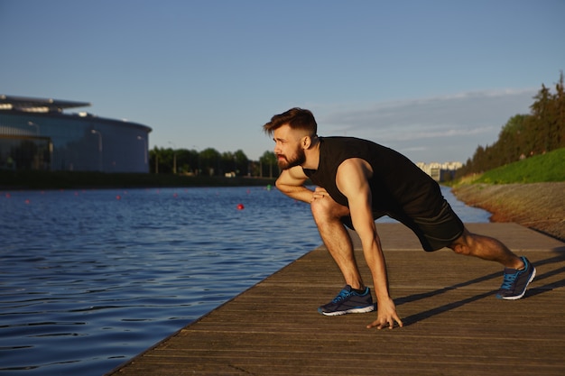 Conceito de exercício físico, atividade, treinamento, esportes e fitness. foto horizontal de um jovem elegante e bonito com a barba por fazer, vestindo roupas esportivas e tênis de corrida, dando mergulhos ao ar livre pela manhã