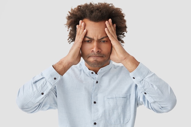 Conceito de expressões faciais negativas. homem afro-americano estressado mantém as mãos na cabeça, pressiona os lábios e olha desesperadamente para baixo, vestido com uma camisa elegante, está em pânico, sofre de dor de cabeça