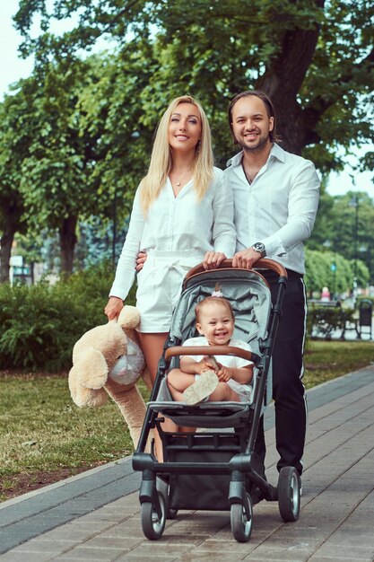 Conceito de família jovem feliz. Linda mãe loira e pai elegante barbudo andando no parque com sua filha em um carrinho de bebê.