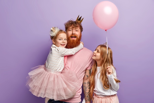 Conceito de família, paternidade e feriados. pai engraçado entretém as crianças, carrega a filha pequena nas mãos, decore o quarto antes da festa, use roupas festivas, isolado sobre a parede roxa.