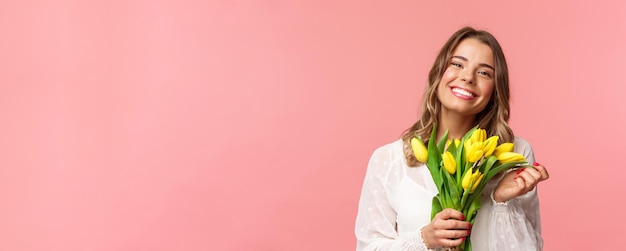 Conceito de felicidade e celebração da primavera closeup de linda jovem loira feliz no vestido branco sonhando acordado após o encontro perfeito segurando tulipas amarelas e sorrindo para a câmera fundo rosa
