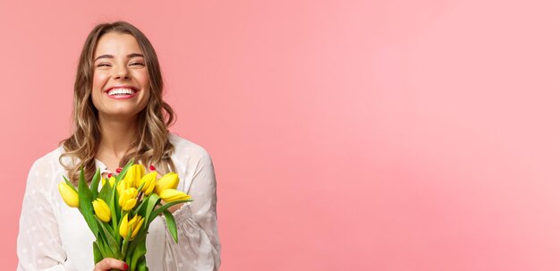 Foto grátis conceito de felicidade e celebração da primavera closeup de menina europeia loira feliz e despreocupada recebe lindo buquê de flores segurando tulipas amarelas sorrindo e rindo fundo rosa