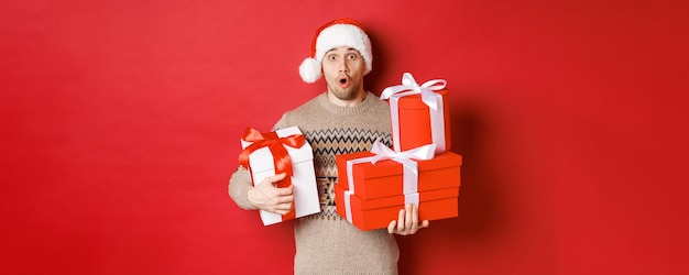Conceito de férias de inverno, ano novo e celebração. Imagem de cara bonito surpreso na camisola e chapéu de Papai Noel, recebendo presentes, segurando caixas com presentes e parecendo espantado.
