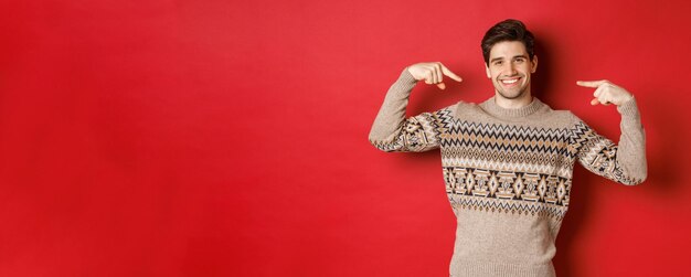 Conceito de férias de inverno de celebração de natal e estilo de vida Imagem de homem bonito feliz na camisola de natal apontando para si mesmo e sorrindo sendo secreta santa em pé sobre fundo vermelho