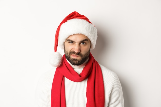 Conceito de férias de inverno e festa de ano novo. Close de cara mal-humorado com chapéu de Papai Noel, carrancudo e fazendo careta, em pé contra um fundo branco