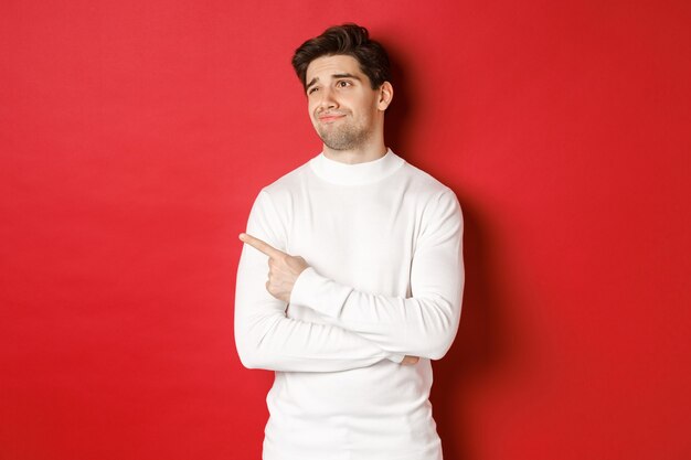 Conceito de férias de inverno. Imagem de um jovem descontente com um suéter branco, olhando e apontando com o dedo esquerdo para algo mediano, cético sobre um fundo vermelho