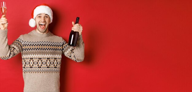 Conceito de férias de inverno, natal e celebração. Animado bonitão de suéter e chapéu de papai noel, aproveitando a festa de ano novo, levantando vidro e garrafa de champanhe, se divertindo