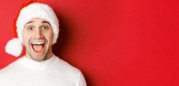 Conceito de férias de inverno natal e celebração Closeup de homem bonito surpreso com chapéu de Papai Noel ouvir incrível oferta promocional de ano novo em pé contra fundo vermelho