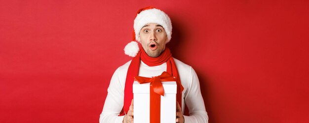 Conceito de férias de inverno, Natal e estilo de vida. Close-up de cara bonito surpreso com chapéu de Papai Noel e lenço, parecendo espantado e segurando o presente de ano novo, em pé sobre um fundo vermelho.