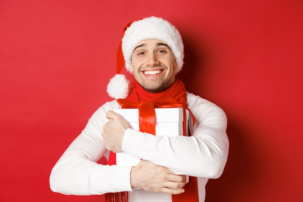 Conceito de férias de inverno, Natal e estilo de vida. Imagem de um cara adorável com chapéu de Papai Noel e lenço, abraçando seu presente de ano novo e sorrindo lisonjeado, em pé sobre um fundo vermelho.