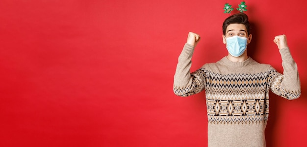 Conceito de férias de natal e coronavírus Homem feliz comemorando o ano novo durante o surto de covid19 usando máscara médica e suéter regozijando-se sobre fundo vermelho