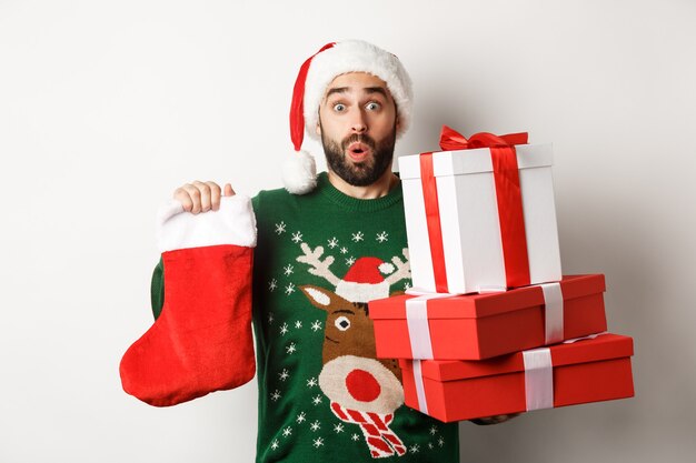 Conceito de férias de Natal e inverno. Homem animado segurando uma meia de Natal e caixas de presente, comemorando o ano novo, trazendo presentes debaixo da árvore, em pé sobre um fundo branco