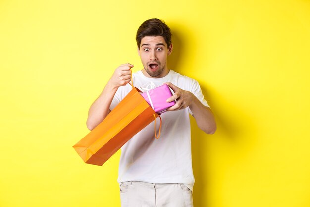 Conceito de férias e celebração. Jovem parecendo surpreso como tirar um presente da sacola de compras, em pé sobre um fundo amarelo.