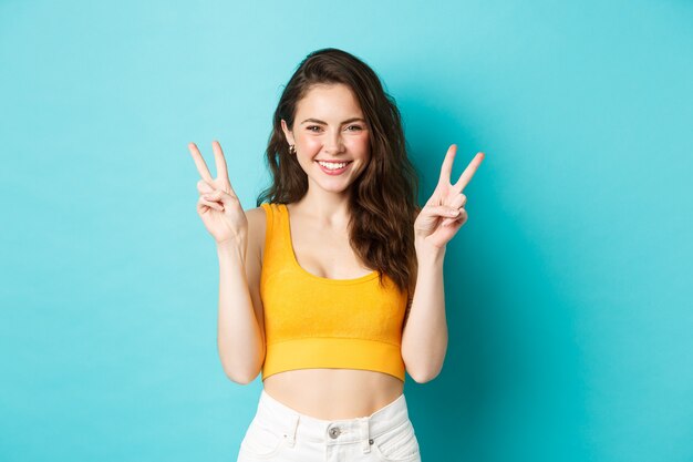 Conceito de férias e emoções de verão. Linda garota coquete sorrindo e rindo, mostrando sinais de V da paz no fundo azul