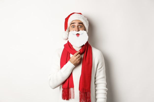 Conceito de festa e celebração de Natal. Jovem engraçado com chapéu de Papai Noel, segurando a máscara de barba branca e fazendo caretas, aproveitando o ano novo, fundo branco.