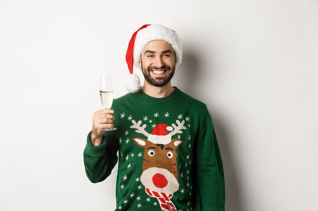 Conceito de festa e feriados de Natal. Bonito homem barbudo com chapéu de Papai Noel e suéter engraçado, bebendo champanhe e comemorando o ano novo. Fundo branco