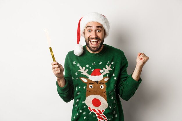 Conceito de festa e feriados de Natal. Bonito homem barbudo regozijando-se, segurando uma festa com fogos de artifício e usando um chapéu de Papai Noel, comemorando o ano novo.