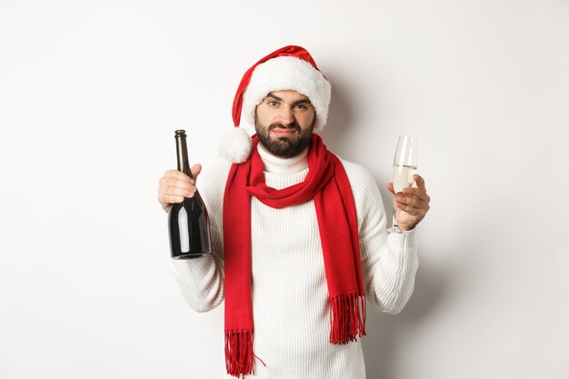 Conceito de festa e feriados de Natal. Homem barbudo cético com chapéu e lenço de Papai Noel, segurando champanhe e reclamando, de pé contra um fundo branco