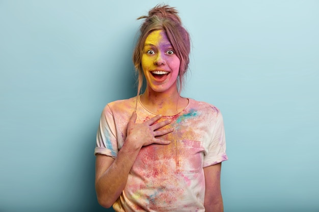 Foto grátis conceito de festival indiano. linda mulher européia impressionada fica de mão no peito, olha de alegria, coberta de tintas coloridas, tem camiseta suja manchada, se diverte com as cores, isolado no azul