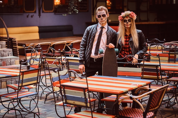 Foto grátis conceito de halloween e muertos. jovem casal atraente com mortos-vivos posando com longboard perto de restaurante mexicano ao ar livre.