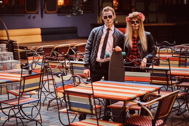 Conceito de Halloween e Muertos. Jovem casal atraente com mortos-vivos posando com longboard perto de restaurante mexicano ao ar livre.