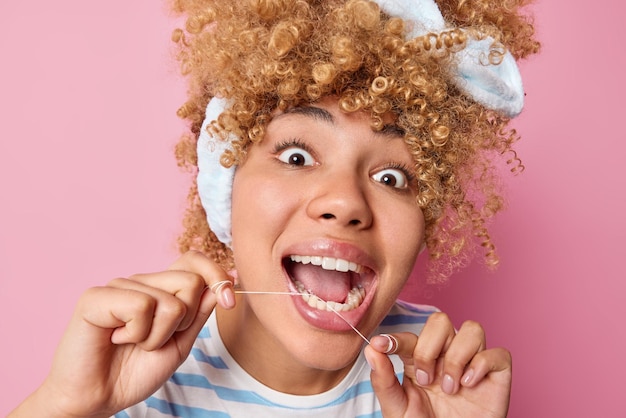 Foto grátis conceito de higiene dental e cuidados com os dentes de estomatologia close-up tiro de jovem de cabelos cacheados usa fio dental limpa os dentes da comida parece surpreendentemente para a câmera isolada sobre fundo rosa