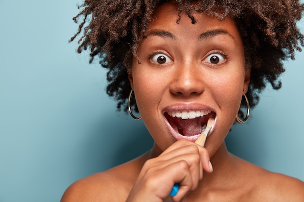 Conceito de higiene e odontologia dental. foto na cabeça de uma jovem afro-americana surpresa com cabelo crespo, usando escova e pasta de dente para limpar os dentes, olhando com olhos esbugalhados isolados no azul