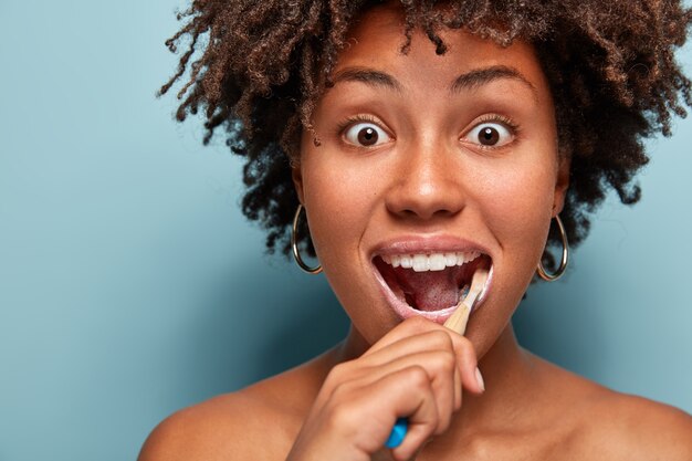 Conceito de higiene e odontologia dental. Foto na cabeça de uma jovem afro-americana surpresa com cabelo crespo, usando escova e pasta de dente para limpar os dentes, olhando com olhos esbugalhados isolados no azul