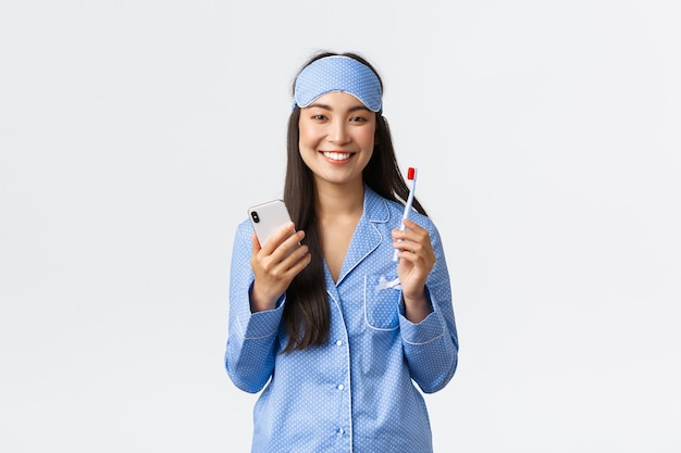 Conceito de higiene, estilo de vida e pessoas em casa. Sorrindo linda garota asiática de pijama azul e máscara de dormir, escovando os dentes antes de dormir e usando o smartphone, mostrando os dentes brancos, fundo branco.