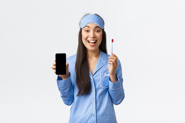Conceito de higiene, tecnologia e pessoas em casa. Sorrindo atraente menina asiática de pijama azul e máscara de dormir mostrando app para ensinar as crianças como usar a escova de dentes e escovar os dentes, segure o smartphone.