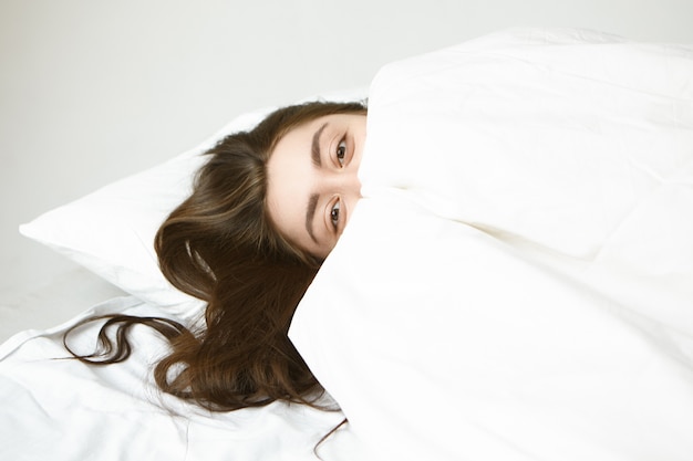 Conceito de hora de dormir, descanso e relaxamento. Mulher jovem e atraente brincalhona com lindos olhos e cabelos escuros relaxando no travesseiro branco, escondendo-se atrás do cobertor, olhando com expressão feliz