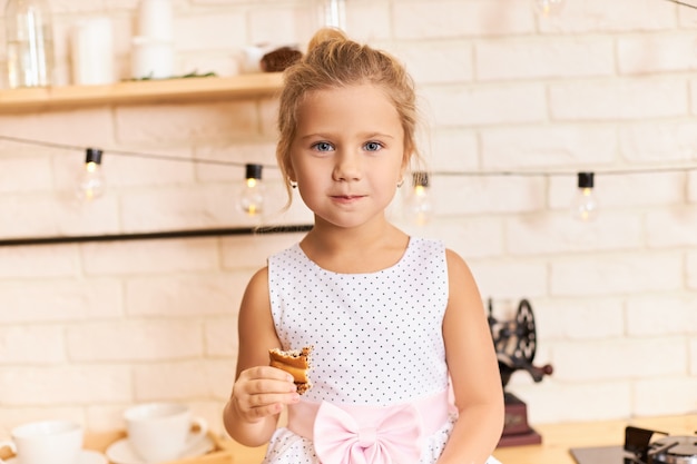 Conceito de infância, diversão e alegria feliz. foto interna da doce adorável menina com um lindo vestido sentada à mesa de jantar no elegante interior da cozinha, rindo, mastigando um biscoito ou torta deliciosos