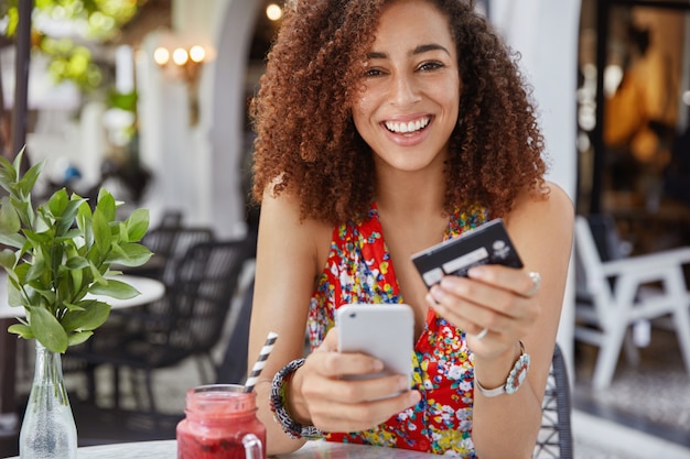 Conceito de Internet banking e comércio eletrônico. Mulher jovem feliz e sorridente com penteado afro, usa um celular moderno e um cartão de crédito para fazer compras online