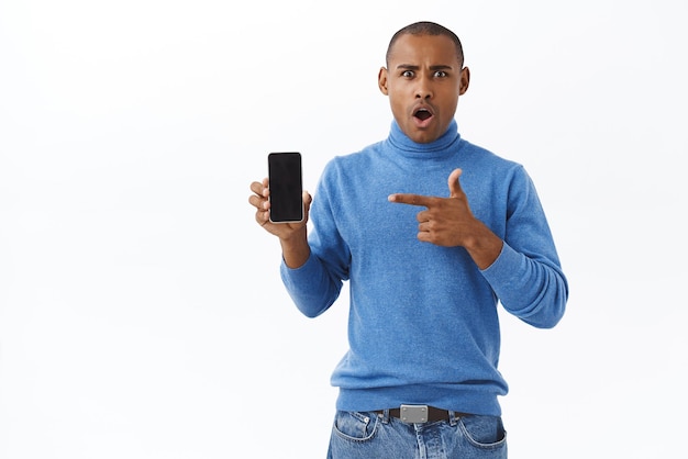 Conceito de internet e pessoas de tecnologia Retrato de homem afro-americano animado chocado mostrando algo na tela do celular com ponto de expressão divertido na tela do smartphone