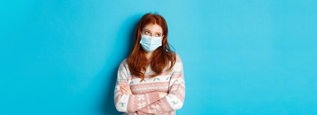 Foto grátis conceito de inverno cobiçoso e pandêmico, adolescente ruiva ignorante com máscara facial revira os olhos e olha para você